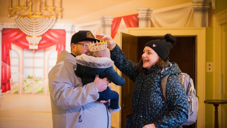 Man and woman holding a baby in the long gallery at Mottisfont, decorated in bright colours and pantomime costumes for Christmas